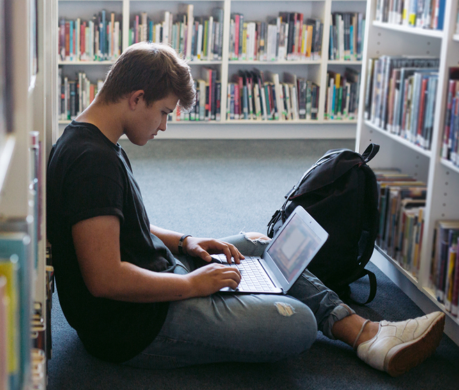 Boy sitting on floor in between library stacks with computer