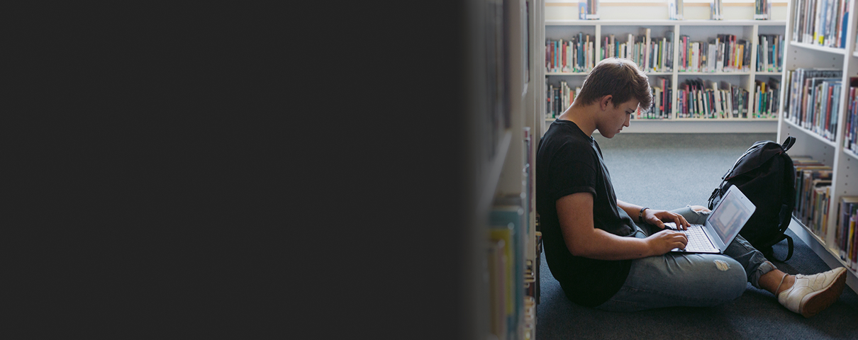 teen on floor in between library stacks looking at computer
