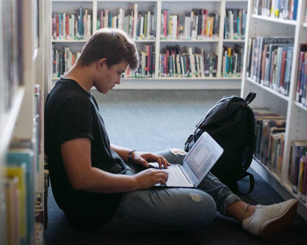 Boy sitting on floor in between library stacks with computer